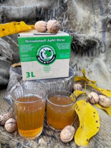 EIn Saftpaket liegt im Hintergrund, zwei Gläser im Vordergrund sind mit Saft gefüllt, die Dekoration besteht aus Walnüssen und Blättern